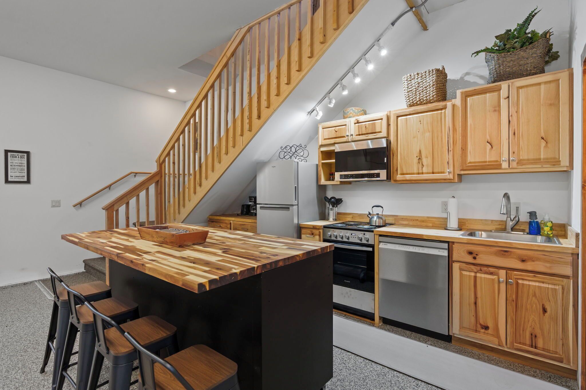 A modern kitchen with wooden cabinets, a central island, bar stools, a wooden staircase, and a stainless steel oven.