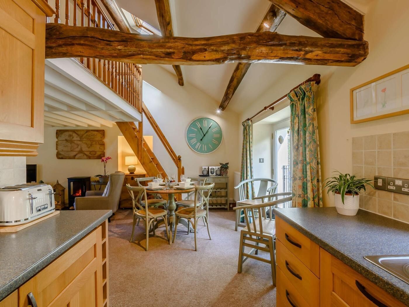 A kitchen with wooden beams, a clock, a dining area with chairs, and a staircase.