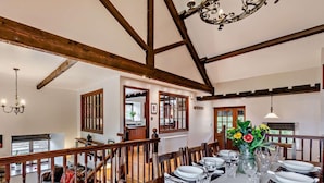 Un comedor con una mesa de madera dispuesta para seis personas, una araña y vista a la cocina a través de un vidrio.