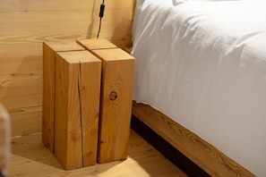 Una mesita de noche de madera con un enchufe, una cama con ropa blanca y un suelo de madera.