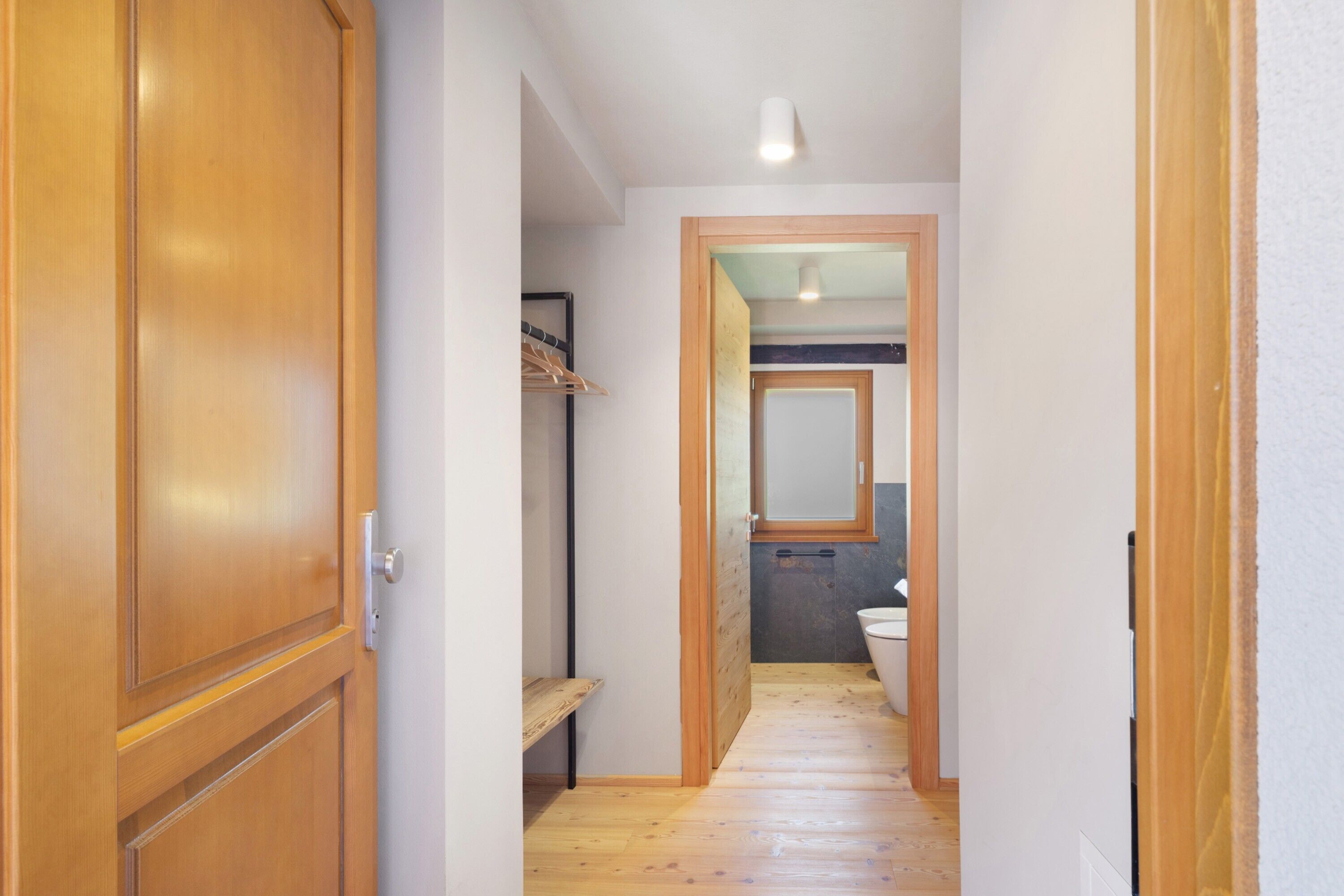 Ein enger Flur mit Holzboden, eine Holztür links und im Hintergrund ein Badezimmer mit Toilette und Spiegel.