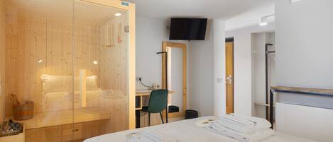 Une chambre d’hôtel moderne dotée d’un sauna en bois, d’un lit avec des serviettes soigneusement pliées, d’un bureau avec une chaise et d’un téléviseur à écran plat fixé au mur.