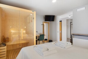 A modern hotel room with a wooden sauna, a bed with neatly folded towels, a desk with a chair, and a flat-screen TV mounted on the wall.