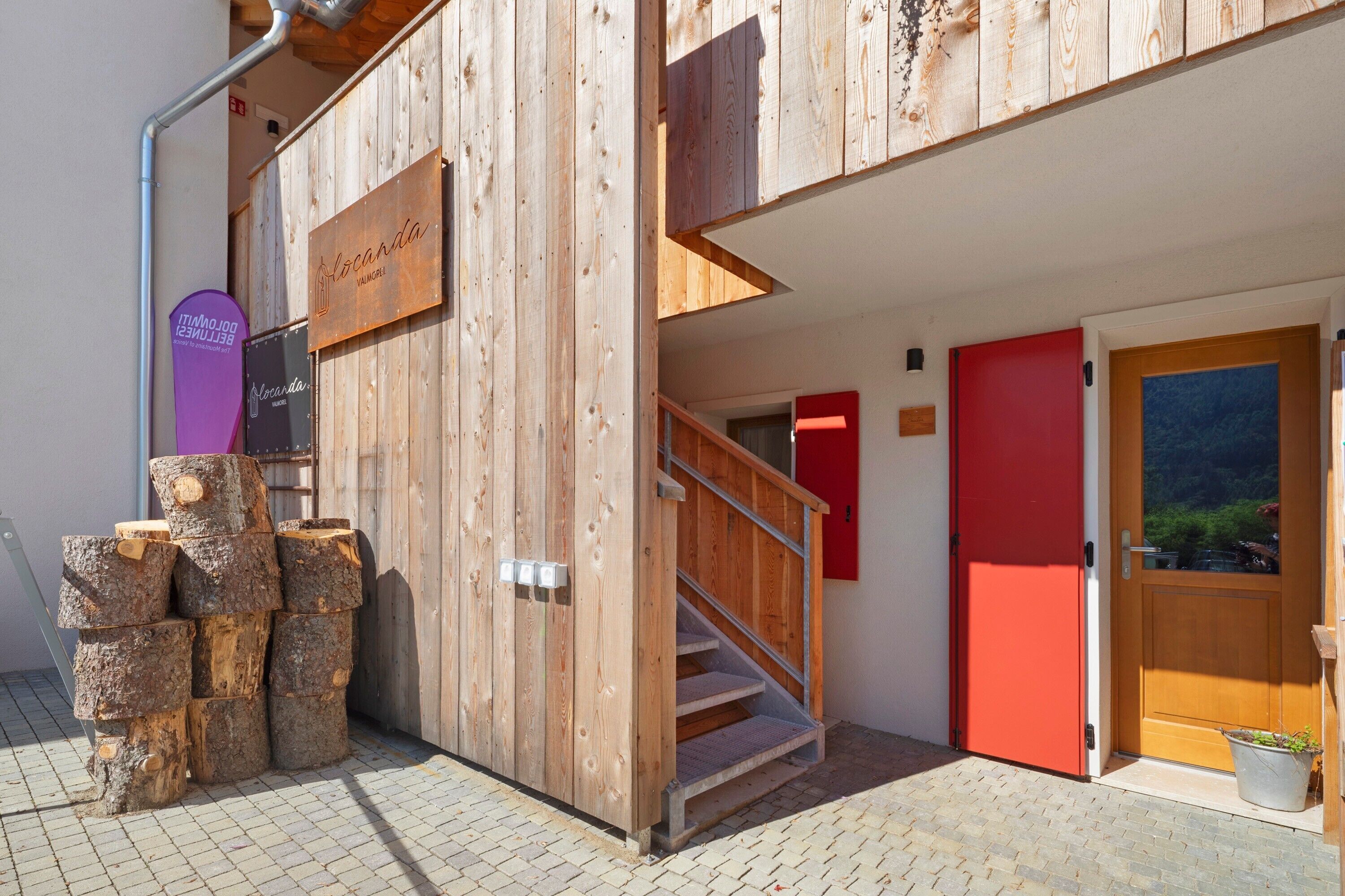 A wooden building entrance with a staircase, a red door, and a signboard.