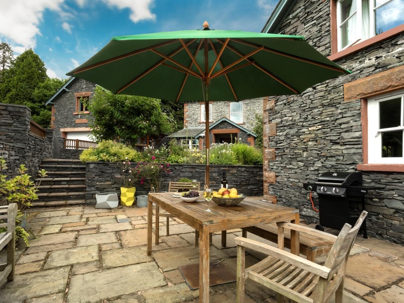 A stone patio with a wooden table and chairs, a green umbrella, and a barbecue grill.
