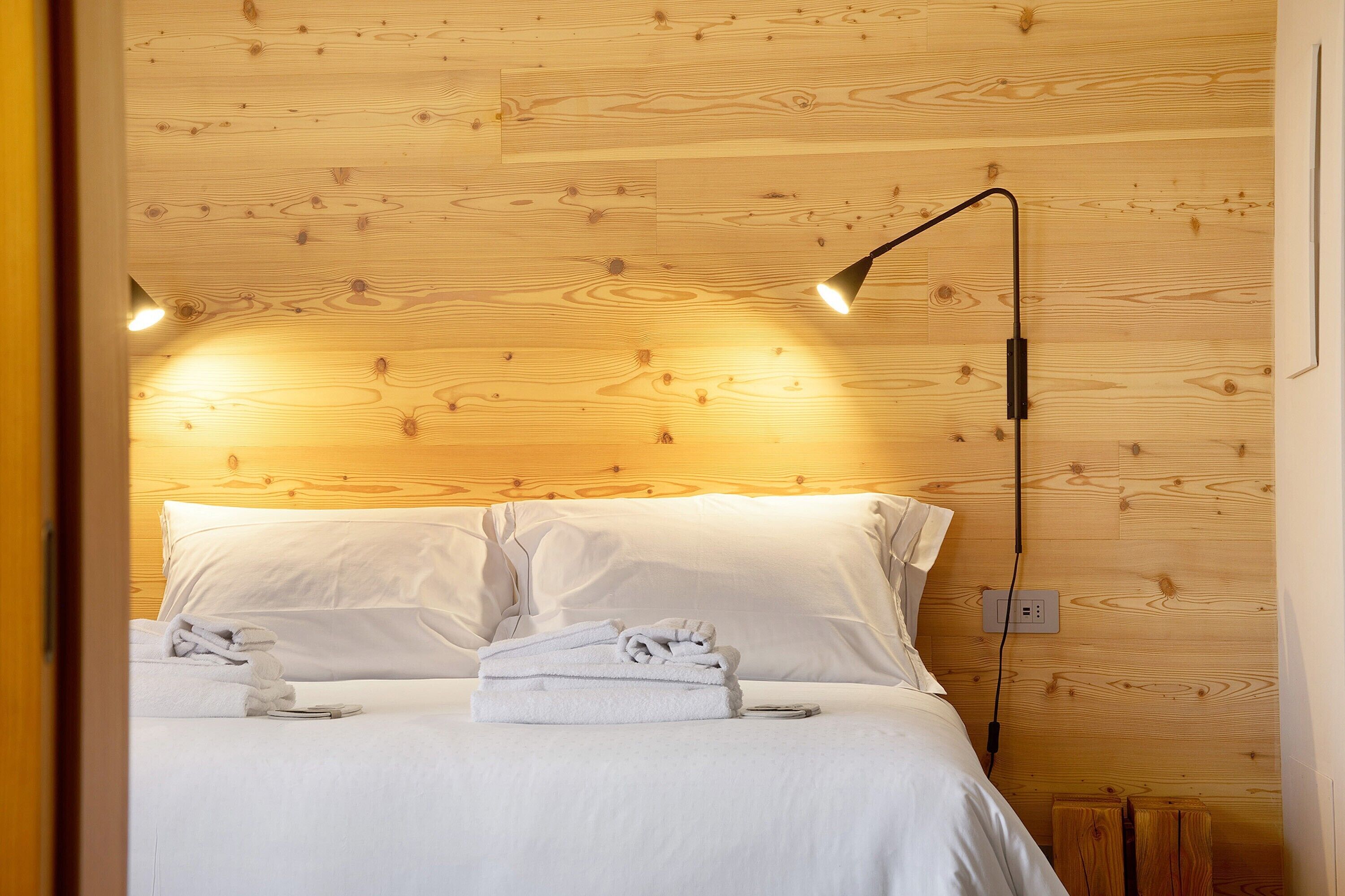 Ein ordentlich bezogenes Bett mit weißen Leinentüchern und zwei aufgerollten Handtüchern, eine Nachttischlampe sowie eine Steckdose an einer Holzwand.