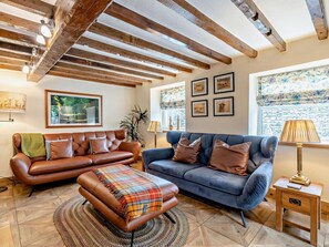 Ein Wohnzimmer mit braunem Ledersofa, einem blauen Sofa, einem gemusterten Hocker, Holzbalken an der Decke, gerahmten Bildern an der Wand und einer Lampe auf einem Holztisch.