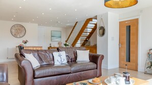 Ein geräumiges Wohnzimmer mit einem braunen Ledersofa, einem Couchtisch mit Gebäck, einer dahinterliegenden Küche und einer Treppe.