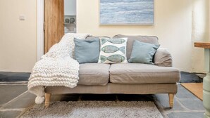 A beige sofa with patterned cushions, a knitted throw, and a framed ocean view painting. - 2 Bed in Llwyngwril (oc-58564) (Llwyngwril)