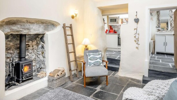 A cozy living room with a stone fireplace, a wooden ladder, a comfortable chair, and a kitchen visible through an open doorway. - 2 Bed in Llwyngwril (oc-58564) (Llwyngwril)