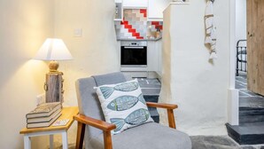 A cozy living room with a chair, a table with books, a lamp, and a kitchen in the background. - 2 Bed in Llwyngwril (oc-58564) (Llwyngwril)