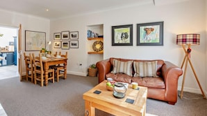 Een woonkamer met een bruine leren bank, een houten salontafel, een eethoek met een houten tafel en stoelen, en kunstwerken in lijsten aan de muren.