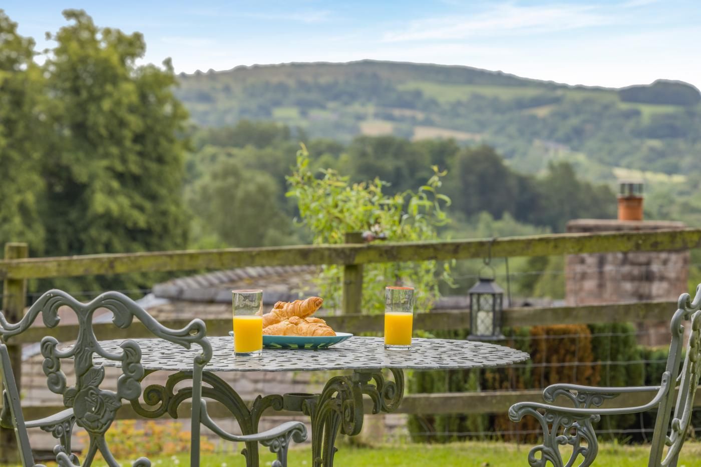 Une table d’extérieur avec un cadre en métal, deux chaises, une assiette de croissants et deux verres de jus d’orange, le tout disposé devant un paysage de collines verdoyantes et un ciel dégagé.