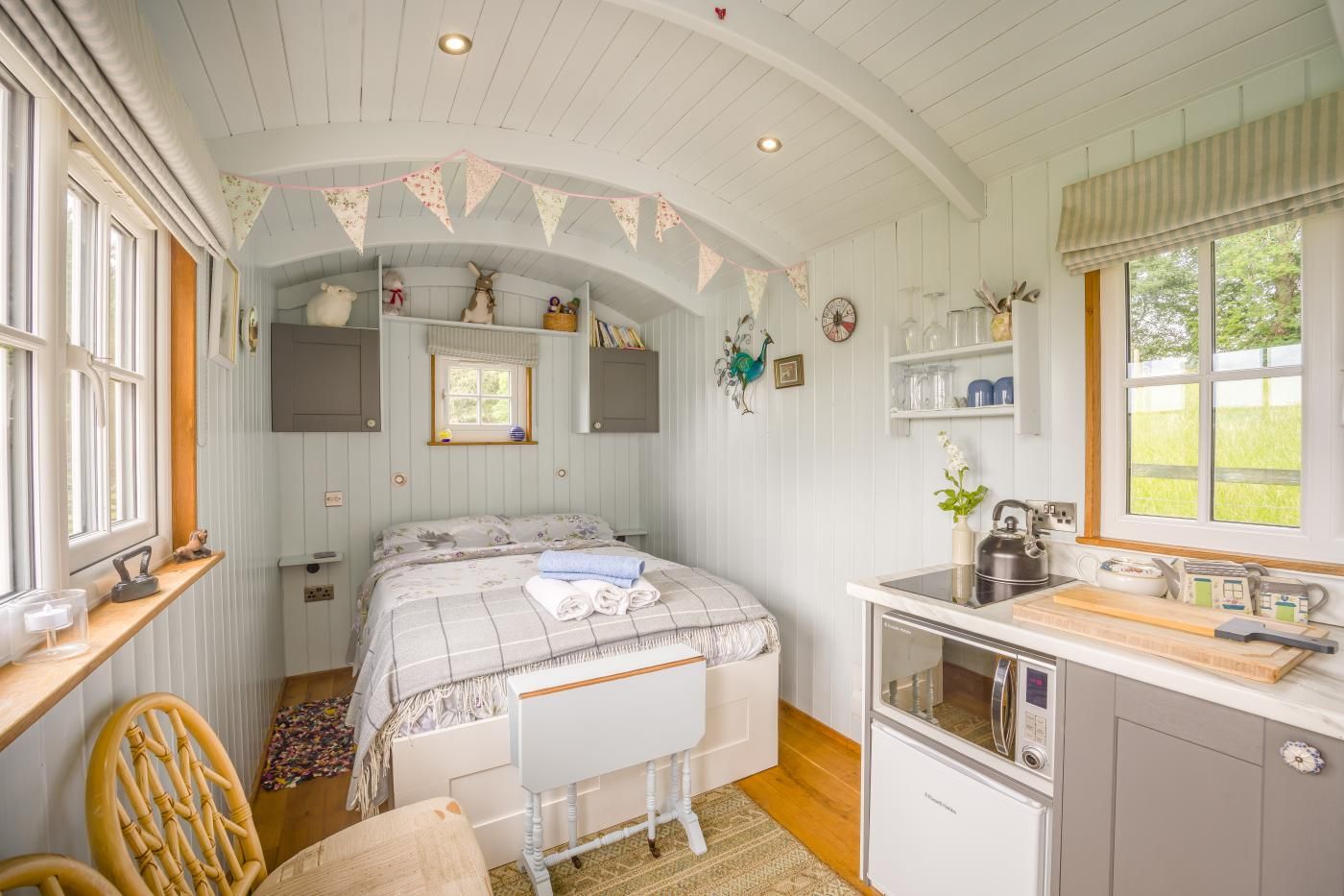 Un dormitorio acogedor con cama, mesita de noche, silla, pequeña cocina con lavamanos y hervidor, y ventana con cortinas.