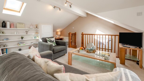 Ein Loft-Wohnzimmer mit schrägem Dach, einem grauen Sofa, einem Couchtisch und einem Fernseher auf einem Holzbodenständer.