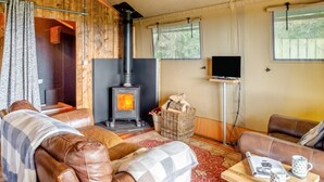 A cozy living room with leather sofas, a wood-burning stove, a television, and a window with a view of trees. - 1 Bed in St. Austell (oc-58376) (Pentewan Valley)