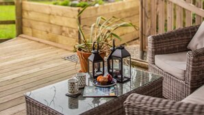 A wicker sofa, a glass coffee table, and two lanterns on a deck with a wooden fence and greenery in the background. - 1 Bed in St. Austell (oc-58376) (Pentewan Valley)