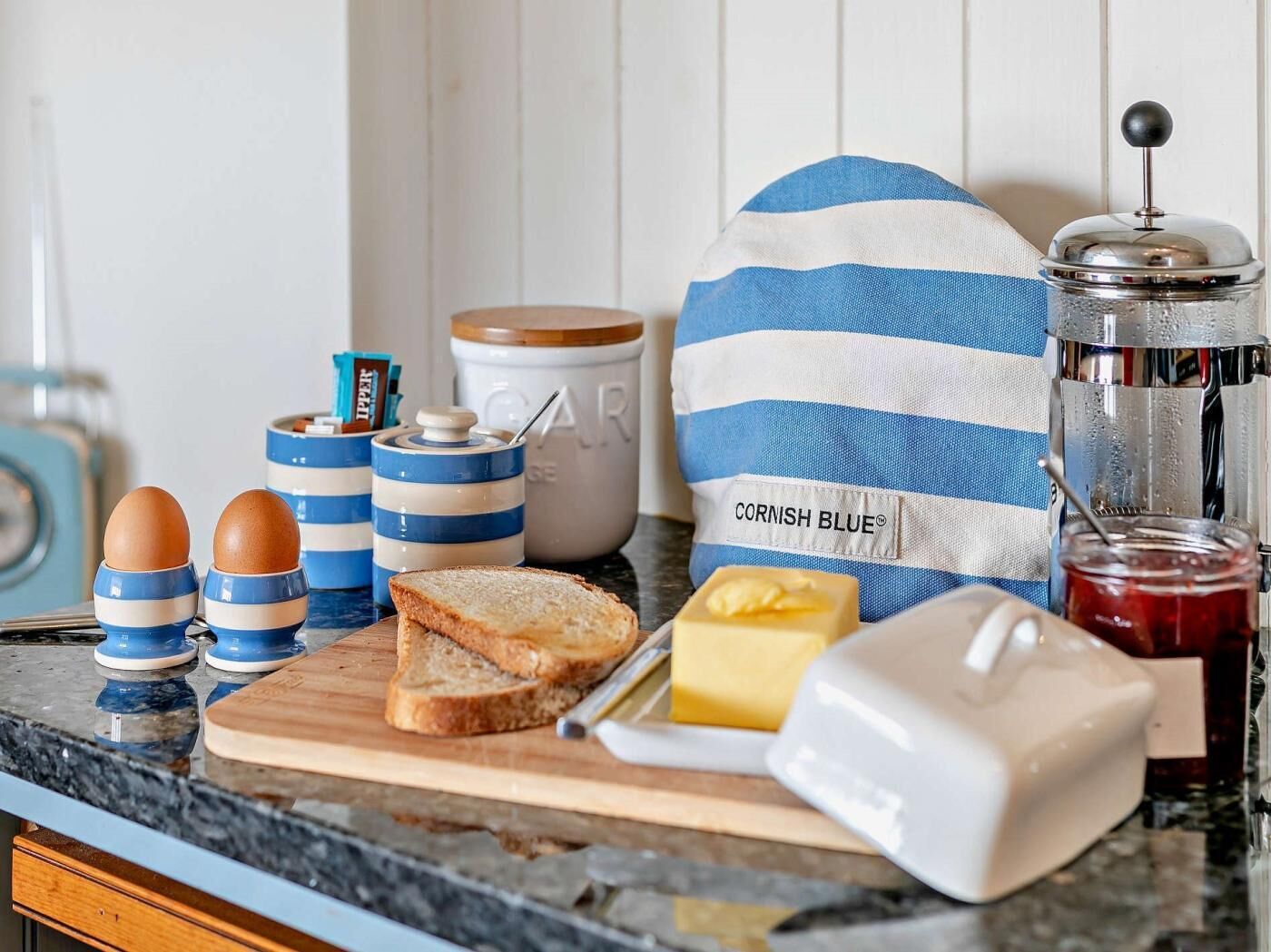 Een aanrecht met gestreepte voorraadpotten, een pot jam, een pot honing, een pot boter, een mes, een snijplank en een zak met de tekst 'CORNISH BLUE'.