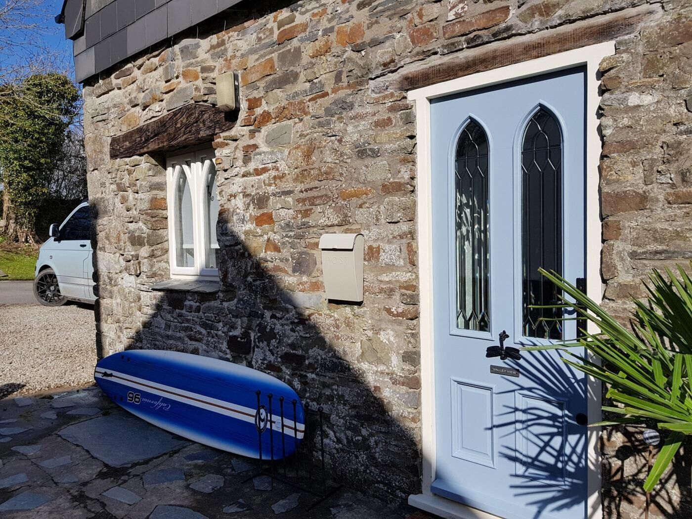 A stone building with a white door, a mailbox, and a blue surfboard leaning against the wall.