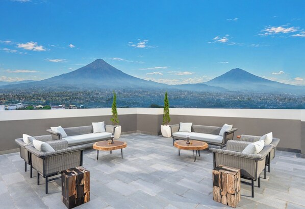 A rooftop terrace with wicker furniture and round tables, overlooking a city and two mountains.