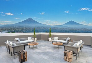 A rooftop terrace with wicker furniture and round tables, overlooking a city and two mountains.