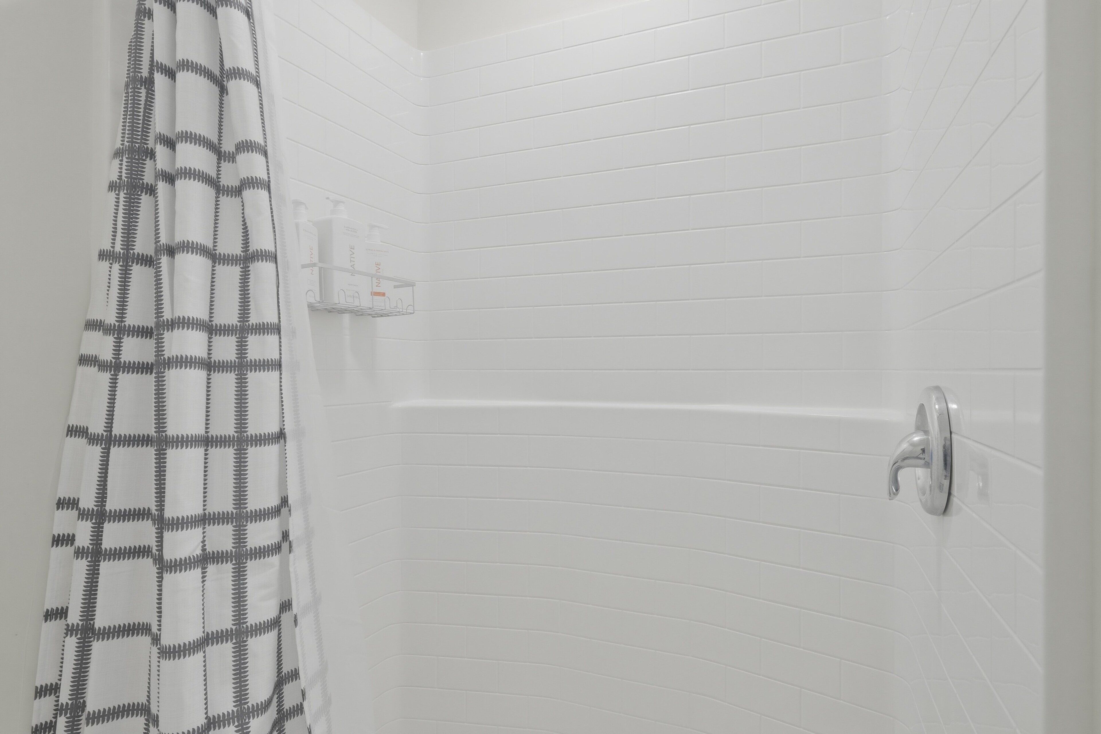 Un baño con paredes de azulejos blancos, una cortina de ducha estampada con un diseño a cuadros y un cabezal de ducha.