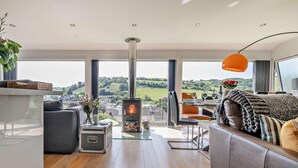 A modern living room with a wood-burning stove, a dining area with a table set for two, and a large window offering a scenic view. - 4 Bed in Torpoint (oc-58602) (Millbrook)