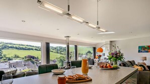 A modern kitchen with a large island, a dining area with a view, and a living space with a sofa and a lamp. - 4 Bed in Torpoint (oc-58602) (Millbrook)