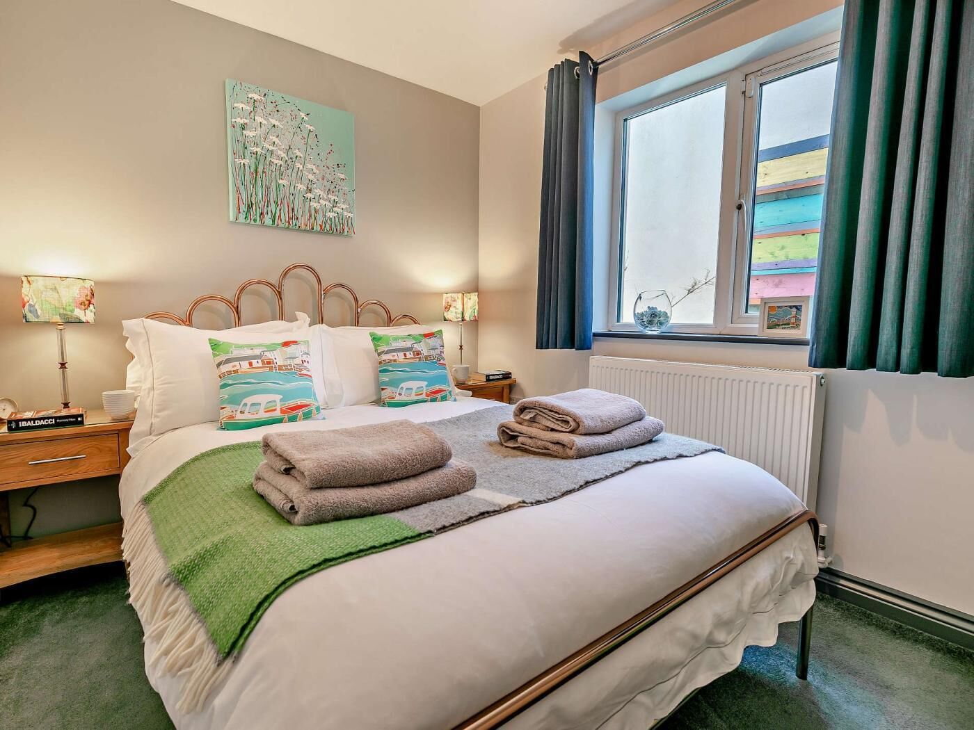 Cama bem arrumada com almofadas, manta verde e toalhas dobradas. Há uma janela com cortinas, um radiador e um quadro na parede.
