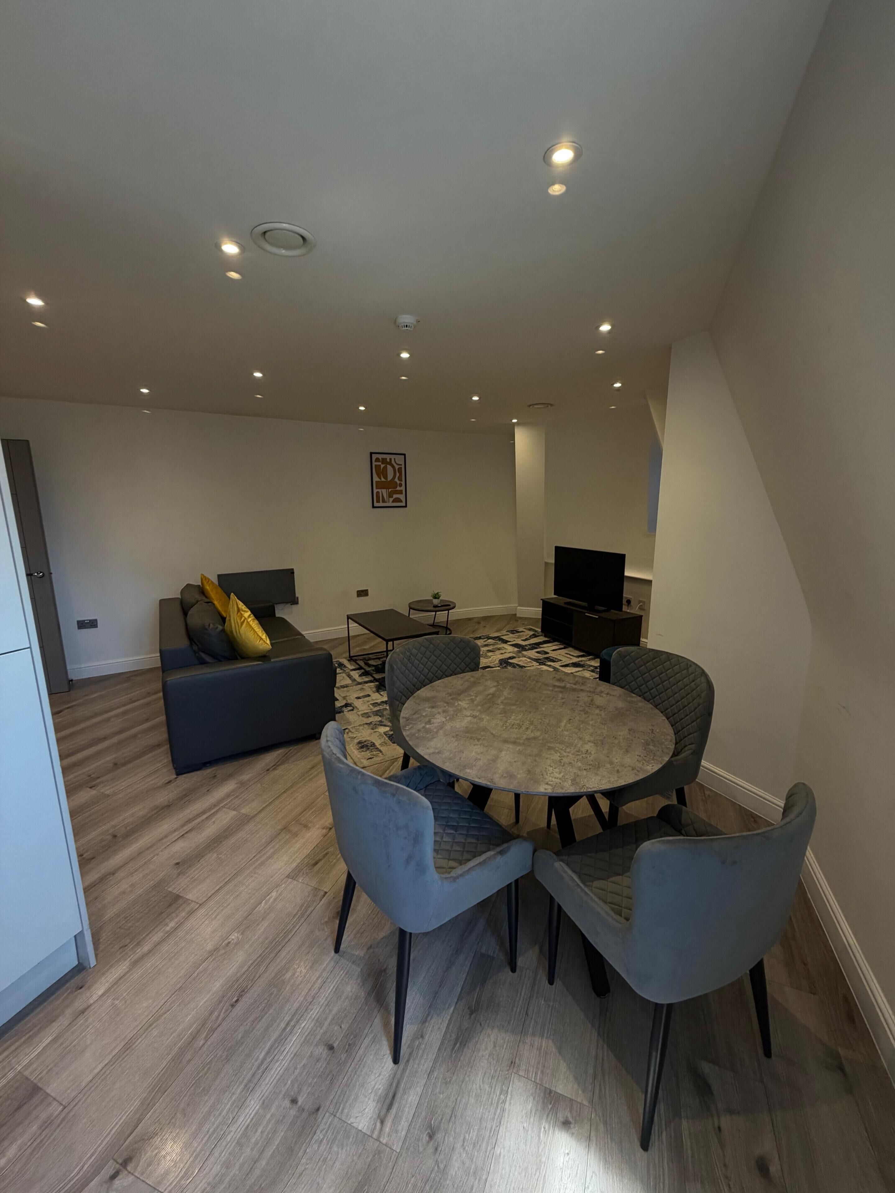 A modern living room with a round dining table, grey upholstered chairs, a dark sofa, and a flat-screen TV.