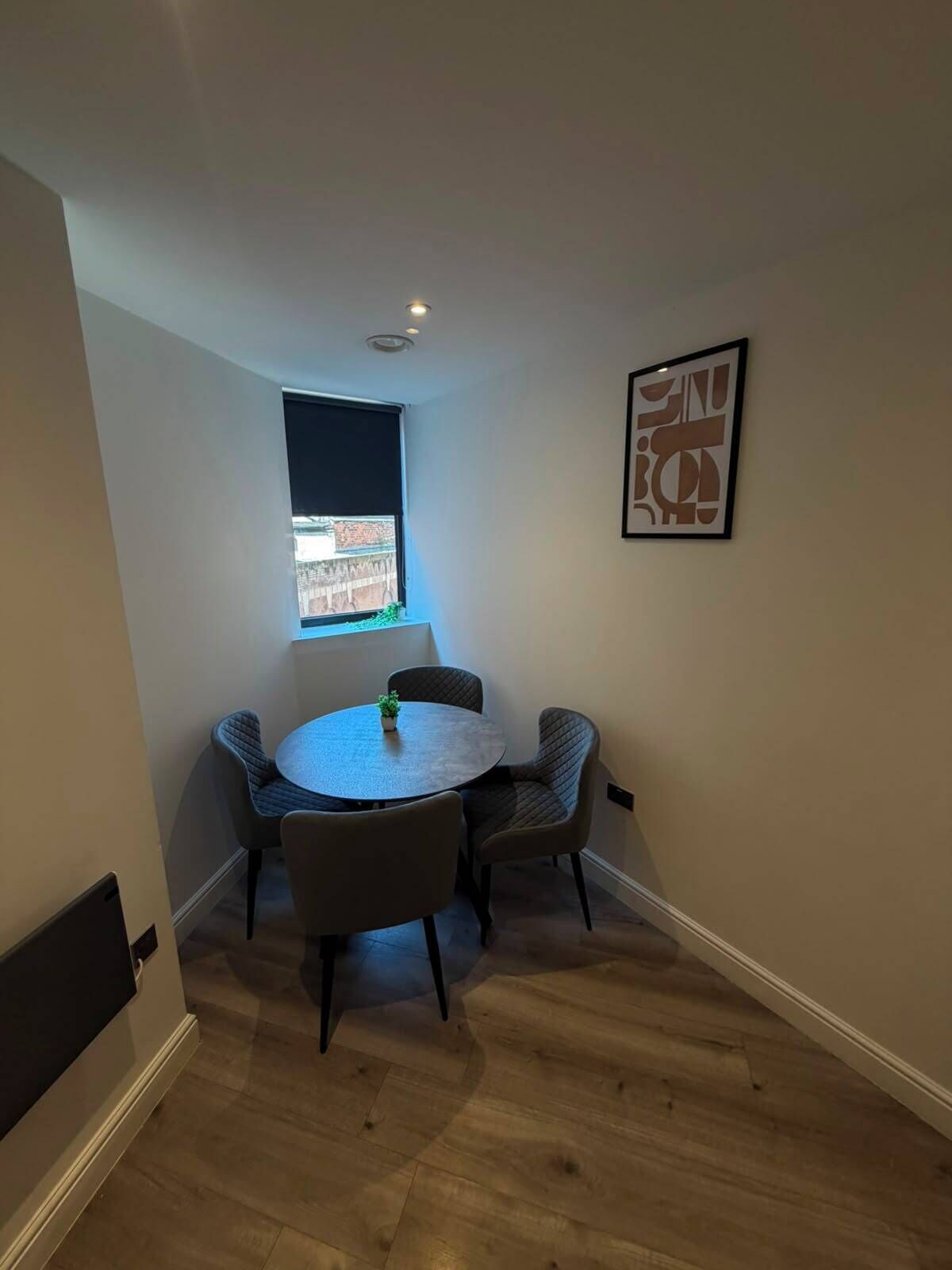 Een kleine eethoek met een ronde tafel en vier stoelen, een raam met uitzicht, en een lijst met een kunstwerk aan de muur.