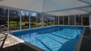 En inomhuspool med väggar av genomskinligt glas, ett tak som skyddar mot väder och vind samt utsikt över en trädgård.