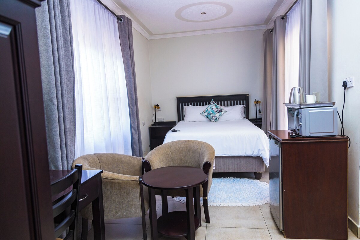 A hotel room with a bed, a desk, two chairs, a small table, a television, and a window with curtains.