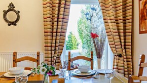 A dining table set for four with chairs, a vase with flowers, and a view of a rocky landscape through curtains. - 3 Bed in Backworth (oc-cn013) (Backworth)