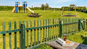 A playground with slides and swings, a picnic table, and a wooden fence. - 3 Bed in Torrington (oc-58822) (Langtree)