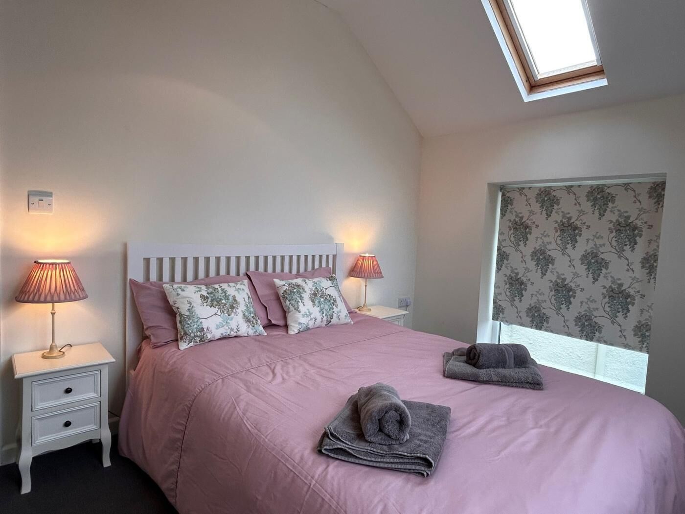 A bedroom with a bed, two bedside lamps, a nightstand, a window with floral curtains, and a skylight.