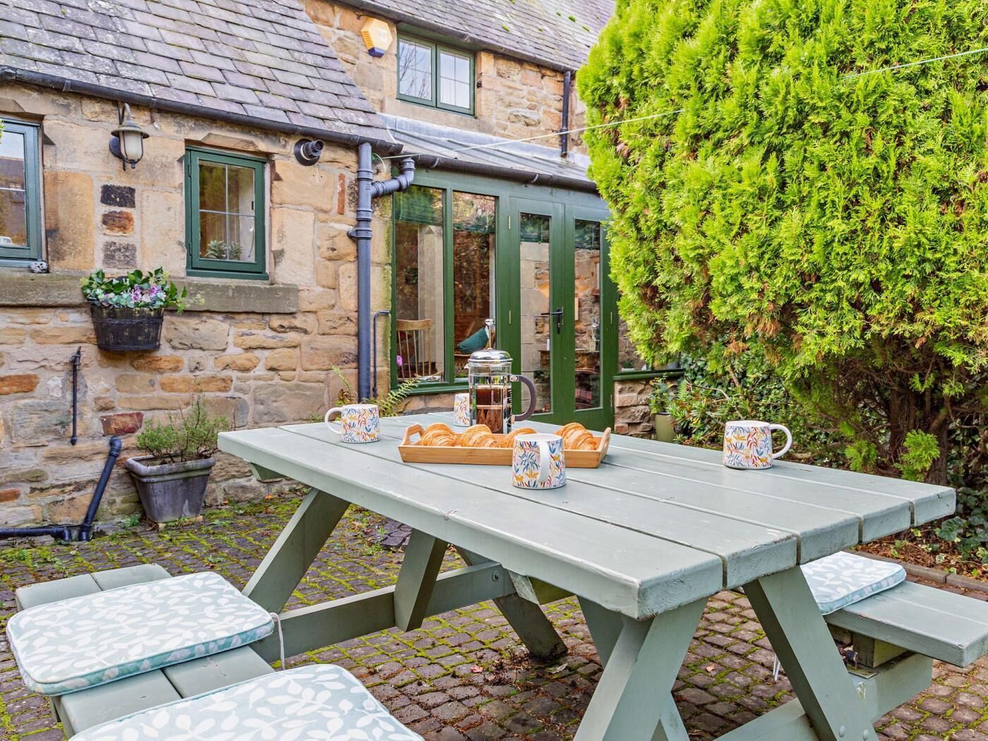 Un bâtiment en pierre avec une porte verte, une table sur laquelle se trouve un plateau de pâtisseries et un banc.