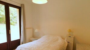 A bedroom with a bed, a bedside table with a lamp, a radiator, and a window with curtains. - Holiday Home 'Villa Moutiques' with Private Terrace, Private Garden and Wi-Fi (Cazaubon)