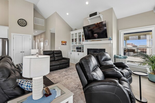 A modern living room with leather sofas, a flat-screen TV, a fireplace, and a view of the outside. - KD2504A-Beautiful, new build home, 5 bedroom, Ocean View, Hot Tub, Elevator! (Kill Devil Hills)