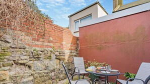 Eine Terrasse mit rundem Tisch und vier Stühlen, eine Ziegelmauer und ein rotes Gebäude im Hintergrund.