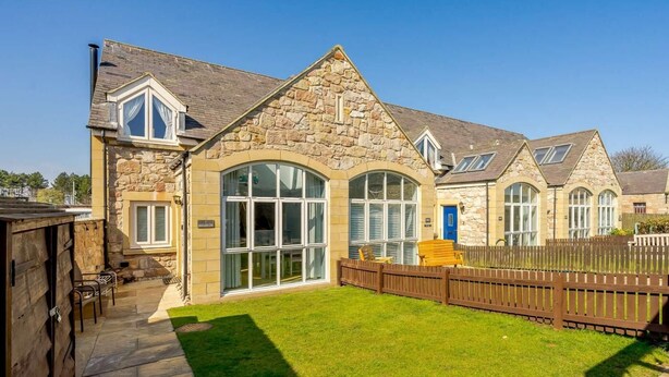 A stone building with a large arched window, a wooden fence, and a well-maintained lawn. - 2 Bed in Beadnell (oc-cn005) (Beadnell)