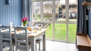 A dining area with a table set for two, a vase of flowers, and a view of the garden through large windows. - 2 Bed in Beadnell (oc-cn005) (Beadnell)