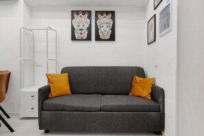 Ein modernes Wohnzimmer mit einem grauen Sofa, zwei senfgelben Kissen, einem weißen Beistelltisch und zwei gerahmten Kunstwerken an der Wand.