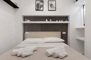 Un llit ben fet amb llençols blancs i dues tovalloles arremolinades al llit. Damunt del llit, hi ha una prestatgeria amb objectes decoratius i una fotografia amb marc.