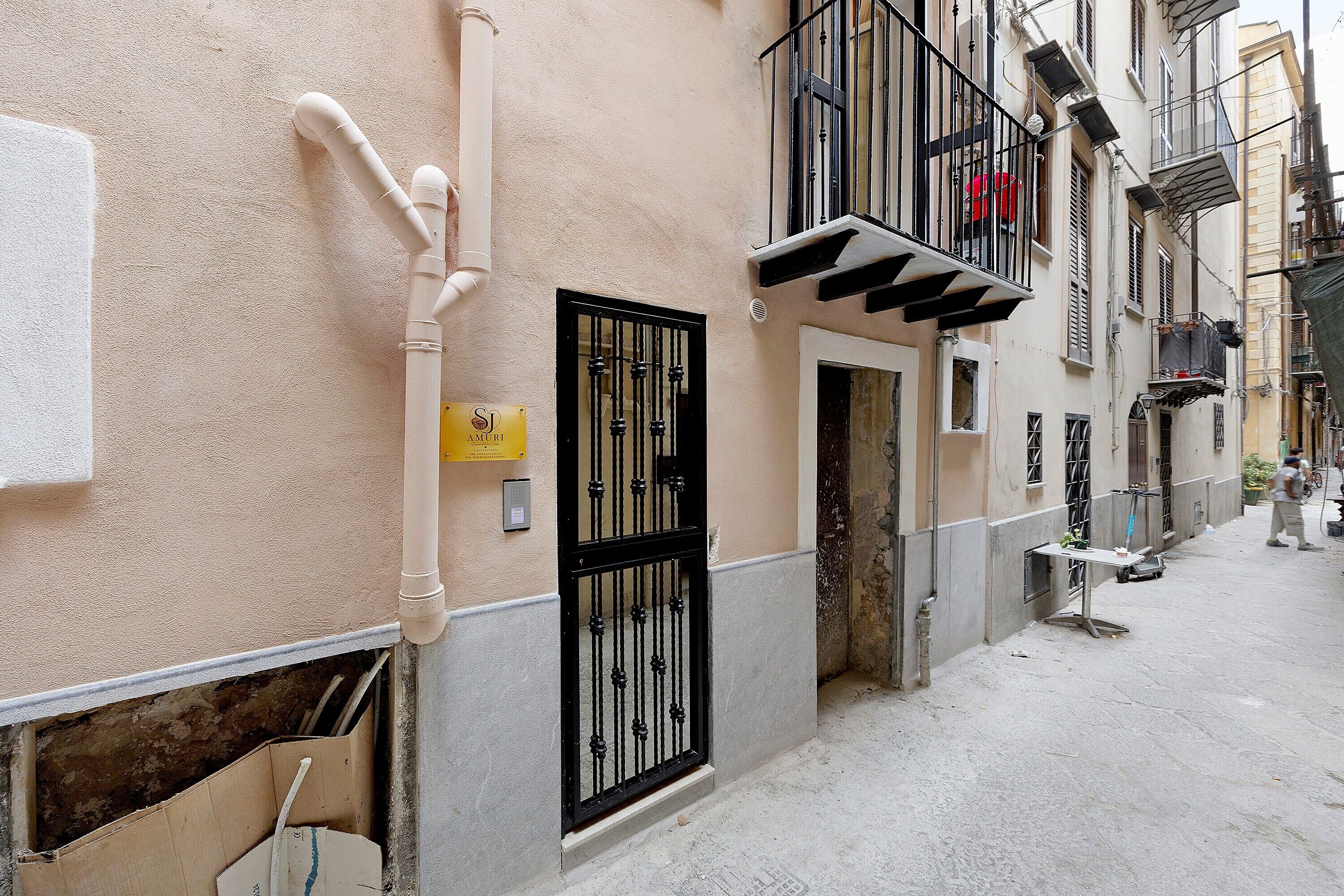Una calle estrecha flanqueada por edificios, una puerta con un portón negro y un letrero amarillo con texto.