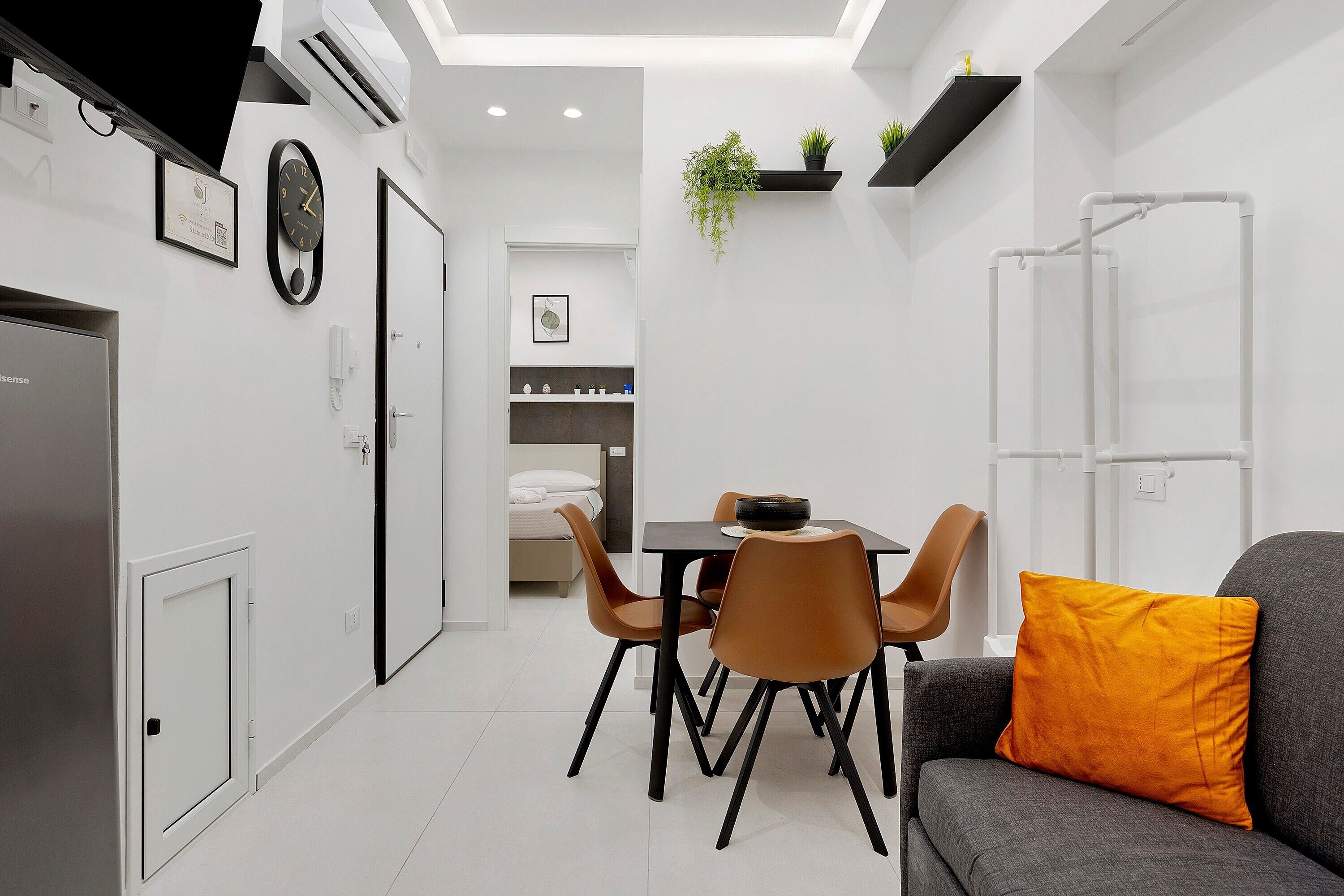 Quarto moderno e bem iluminado com área de jantar, um sofá com almofada laranja e uma cama ao fundo.