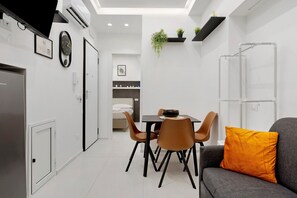 Habitació moderna i ben il·luminada amb zona de menjador, un sofà amb coixí taronja i un llit al fons.