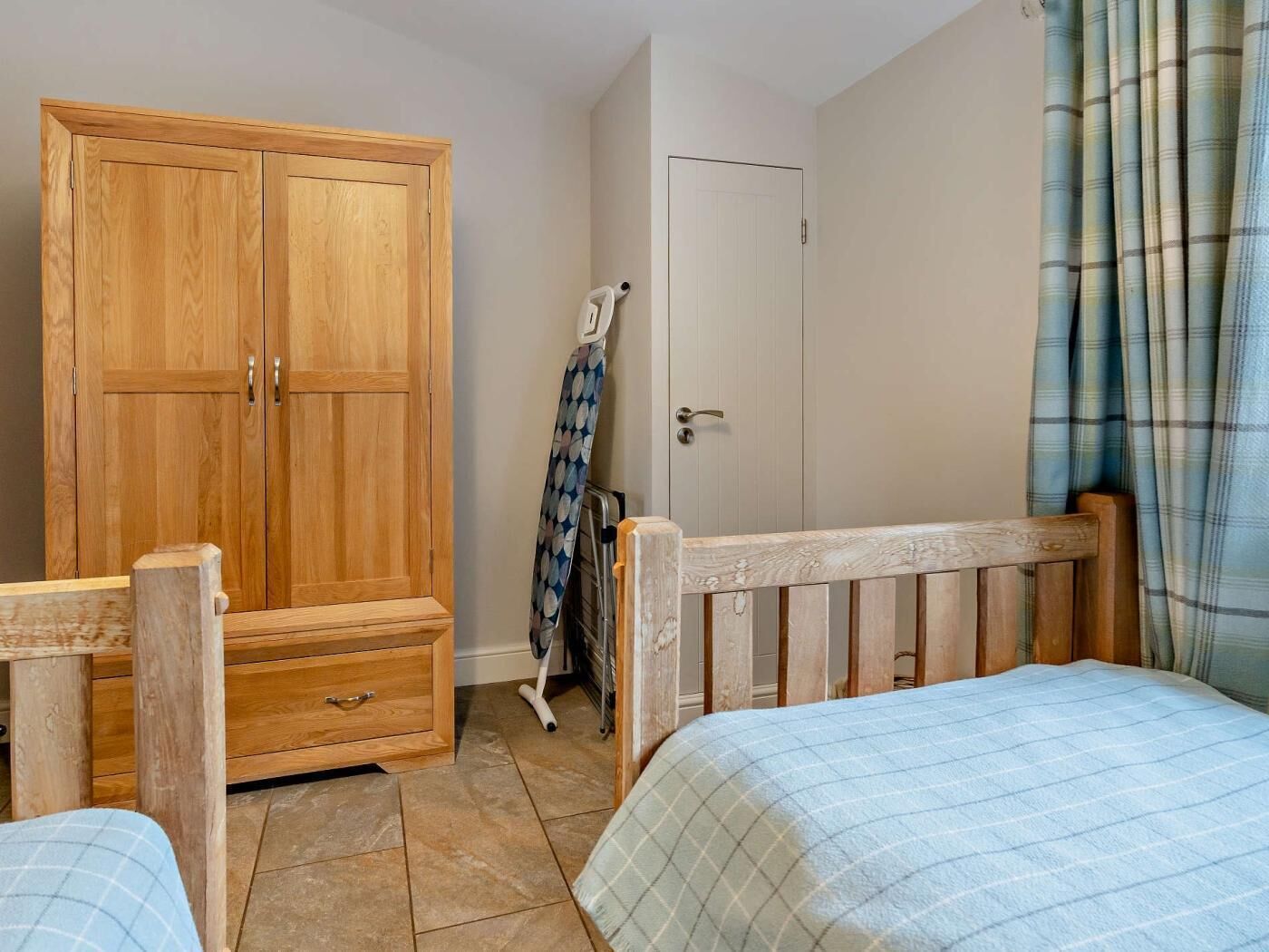 A bedroom with a wooden wardrobe, a bed with a plaid bedspread, a white door, and a window with curtains.