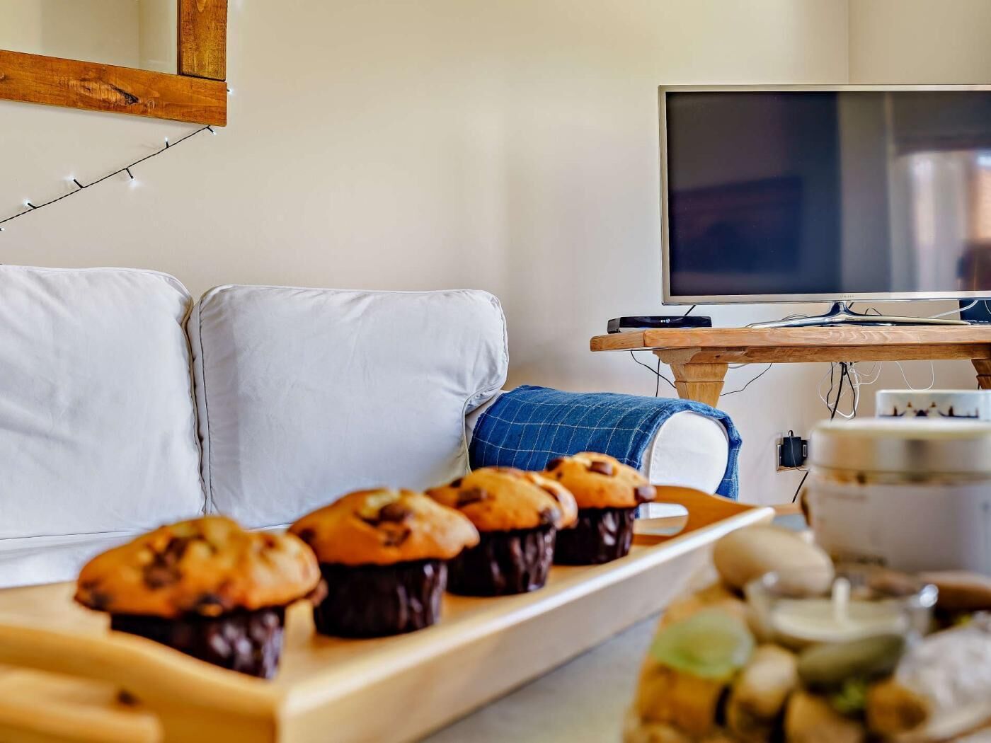 A living room with a sofa, a wooden coffee table with muffins, a television, and a kitchen area with a kettle.