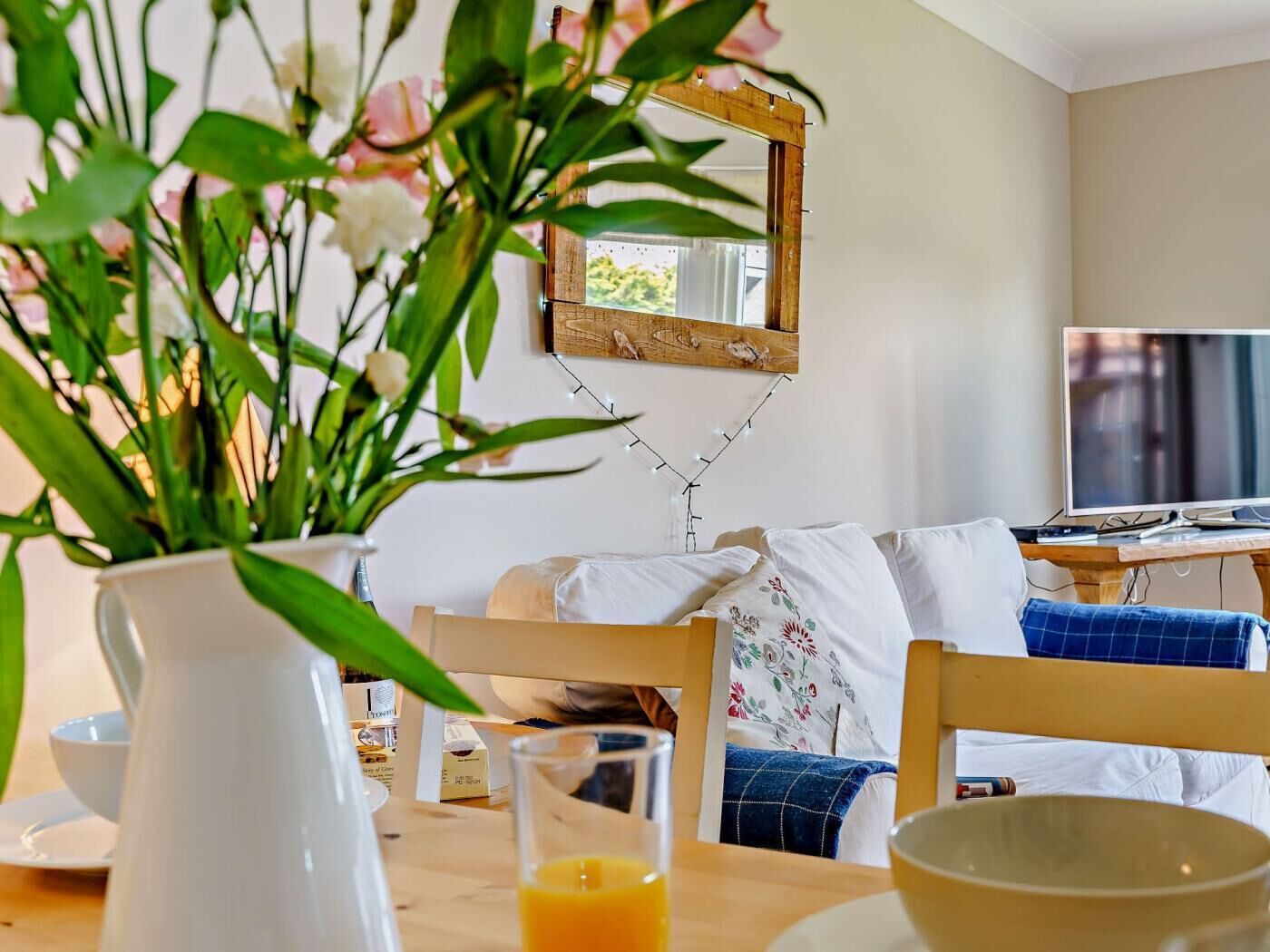 A dining area with a wooden table, chairs, a white vase with flowers, a glass of orange juice, and a television in the background.